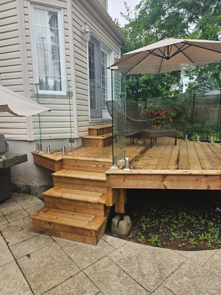 Wooden deck with glass railing attached to a beige house, outdoor seating, and large beige umbrella.