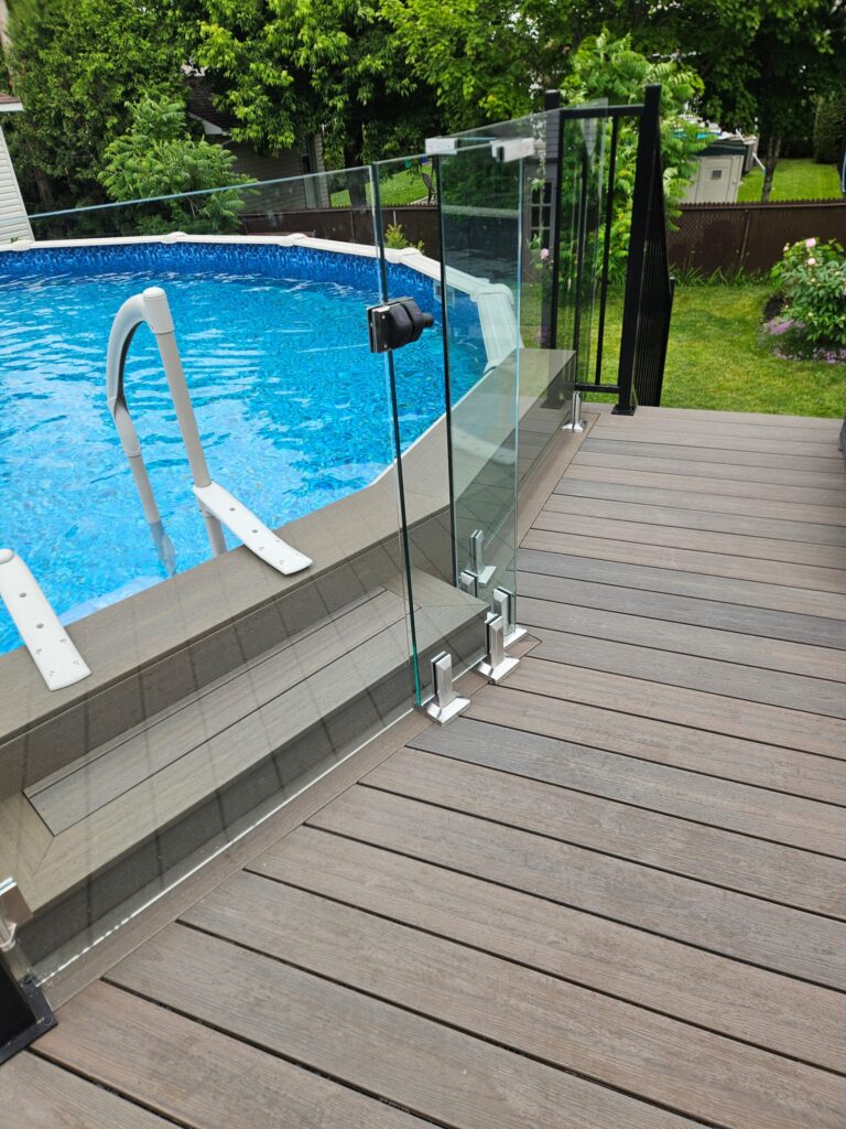 Glass-panel safety railing along a wooden deck surrounds a bright blue backyard pool with metal posts and a ladder emergency step nearby.